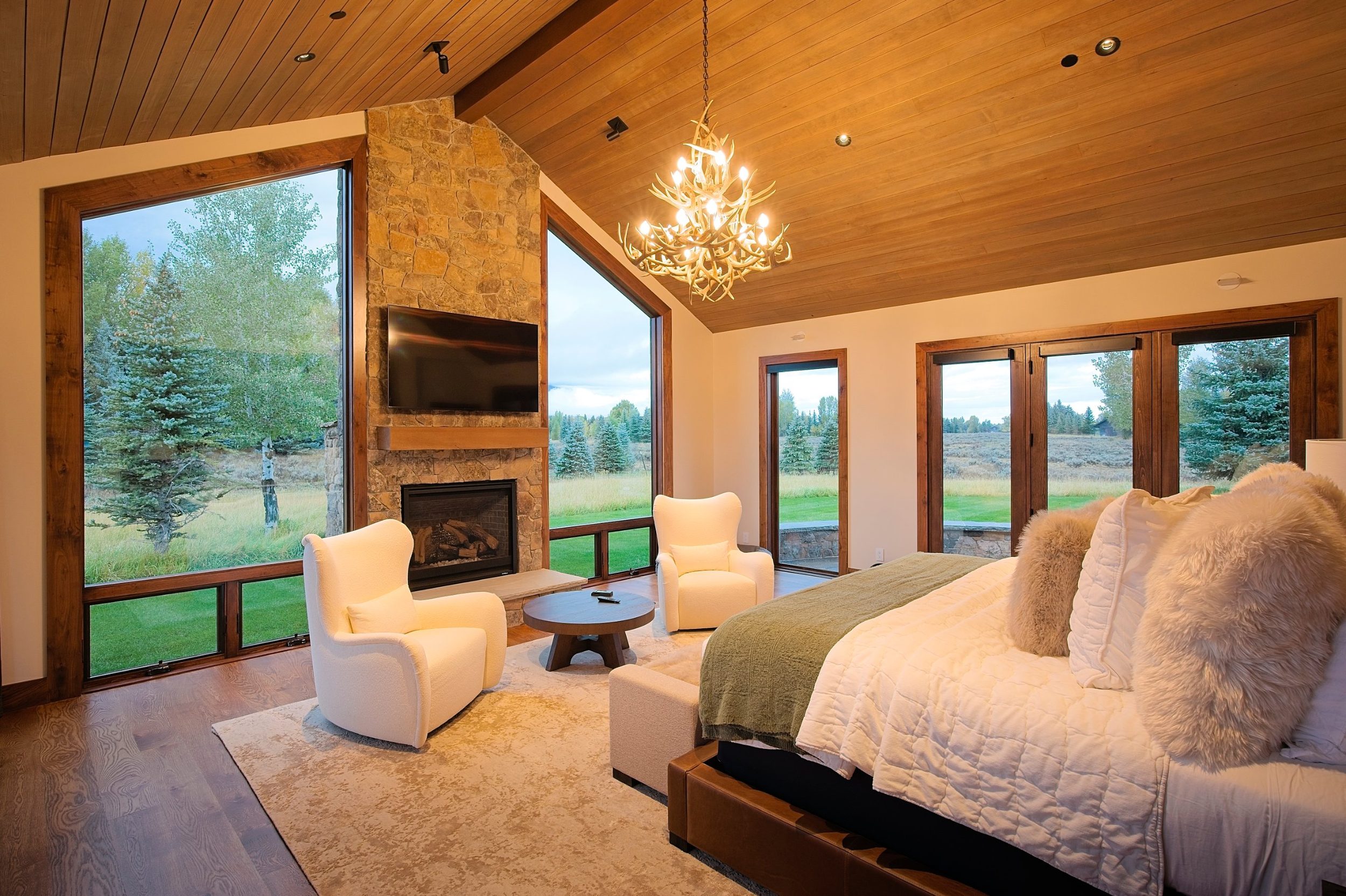 Primary bedroom with stone fireplace, vaulted wood ceiling, and floor-to-ceiling windows to the meadow