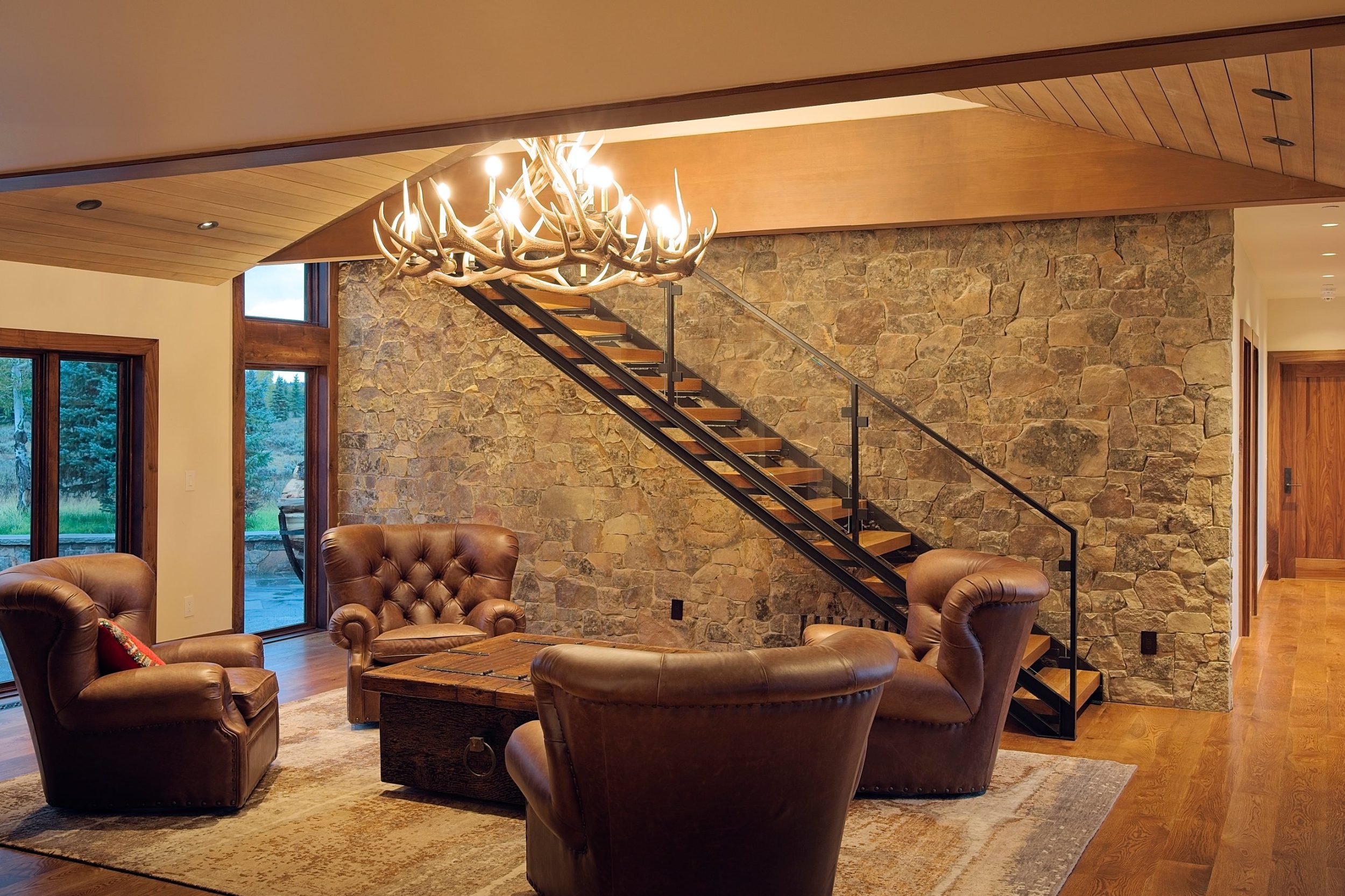 Sitting room with leather chairs, antler chandelier, and open steel stair against a stone wall