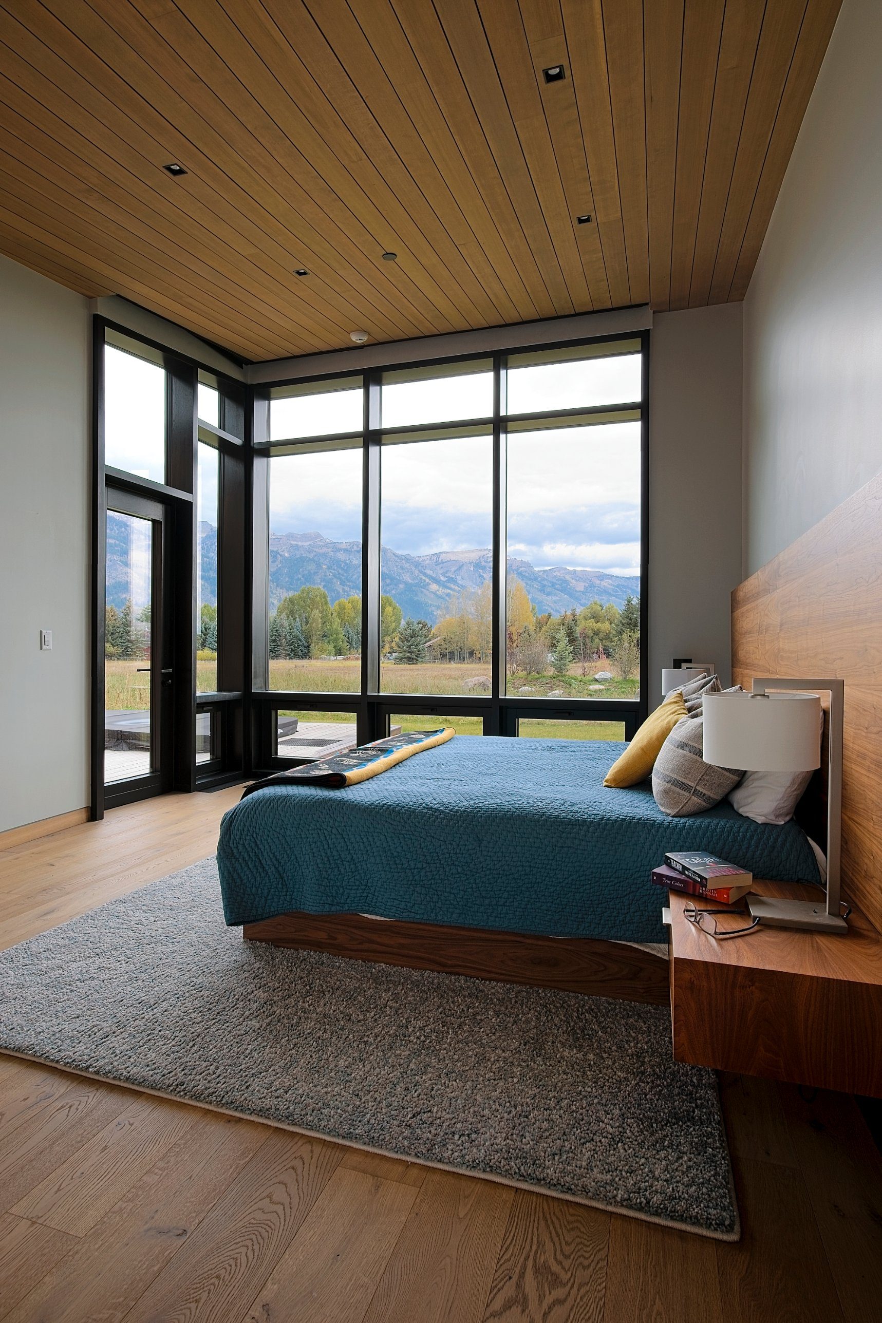 Bedroom with corner floor-to-ceiling windows, wood plank ceiling, and walnut headboard overlooking a meadow