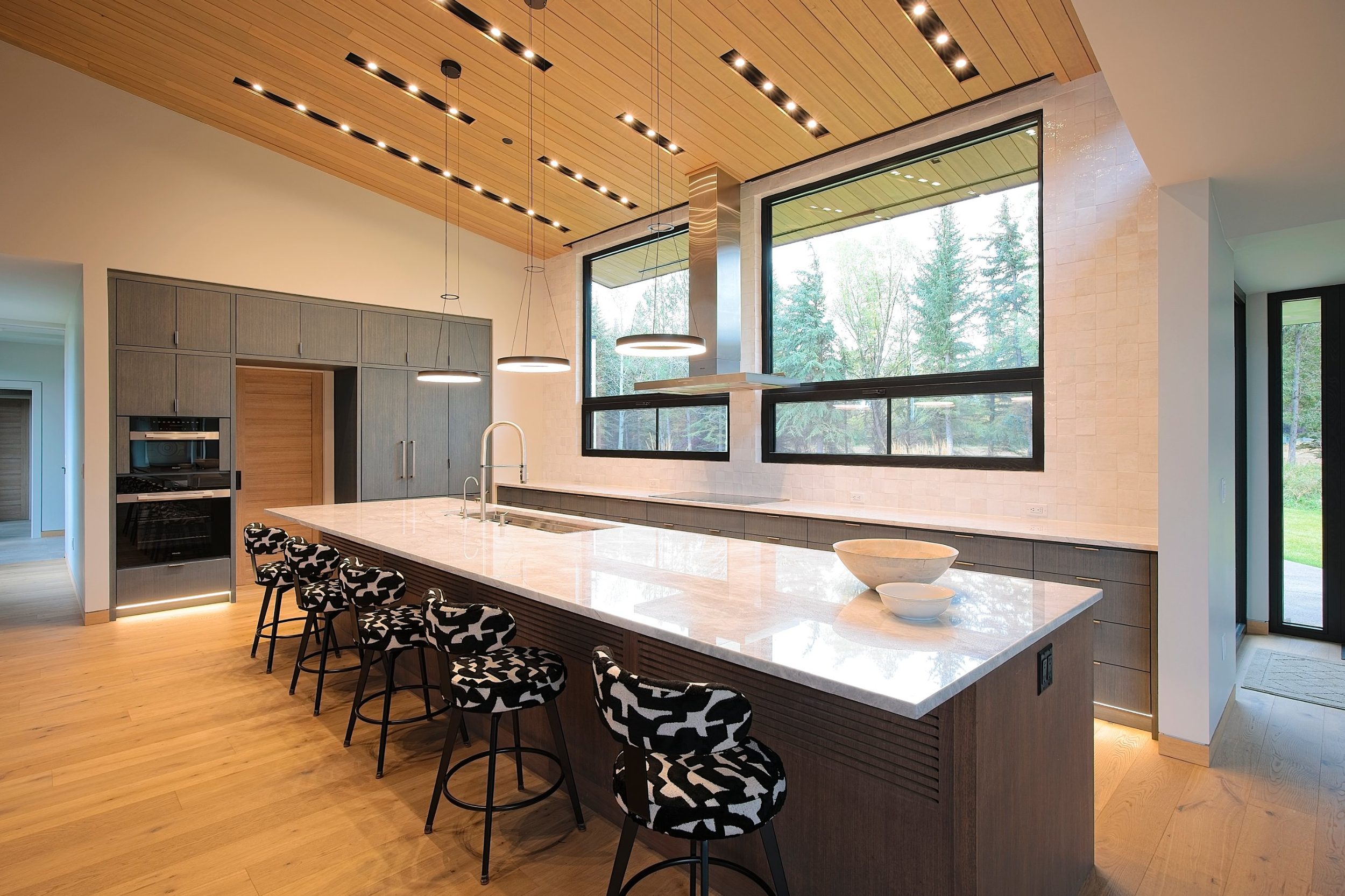 Modern kitchen with long marble island, linear ceiling lighting, and large windows to the trees